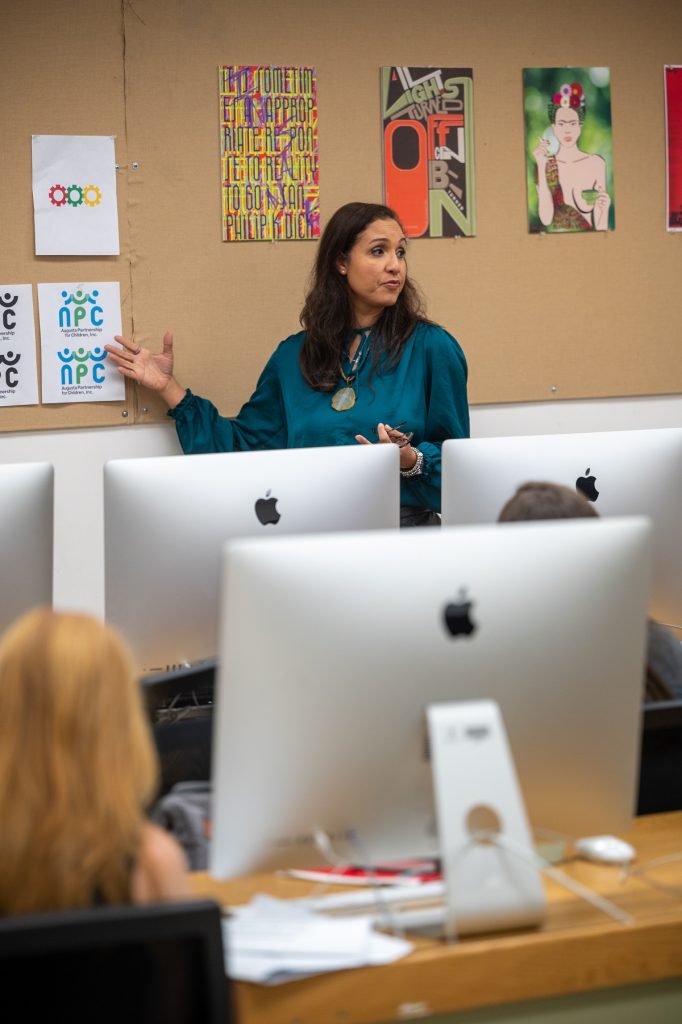 A female college professor teaches a college class on graphic design. She is standing at the front of a classroom and is pointing at several pages on a board that feature different logos from a class project.