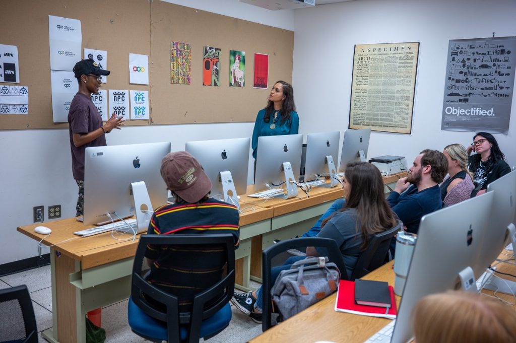 A female college professor interacts with a college student at the front of a classroom as other students look on. The professor and student are discussing logo designs that are pinned to a board between the student and professor.