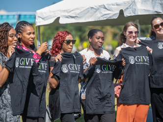 Seven women hold up t-shirts that have the Augusta University funding campaign IGIVE logo on them.