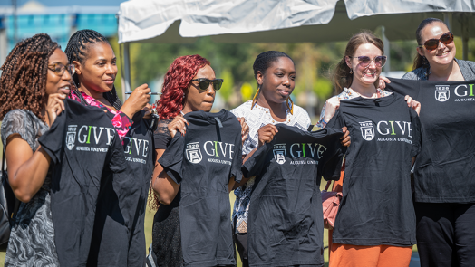 Seven women hold up t-shirts that have the Augusta University funding campaign IGIVE logo on them.