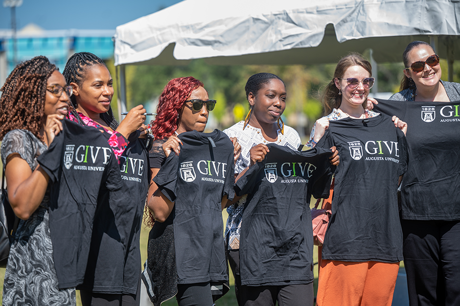 Seven women hold up t-shirts that have the Augusta University funding campaign IGIVE logo on them.