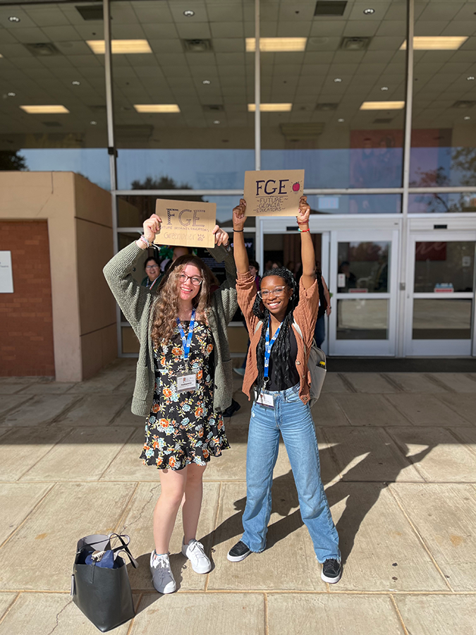 Two female students smile while holding papers that say "FGE" on them over their heads in front of a building.