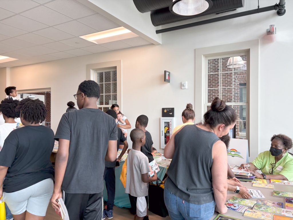 Several families gather around long tables covered in children's books. Parents look at the titles and pick out books for their children during a community event.