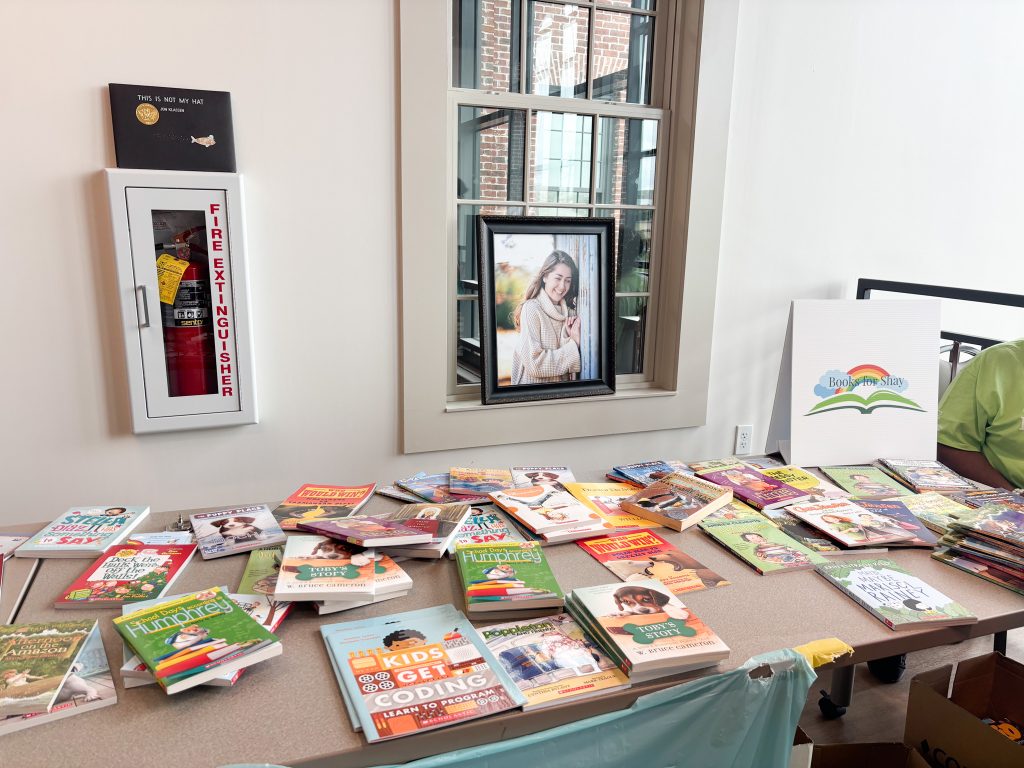 Children's books are spread out over several long tables. There is a sign on one of the tables that reads, "Books for Shay," a program that provides children in underserved areas with books. There is also a photo of a young woman behind the table, celebrating the life of the woman whom the event is named after.