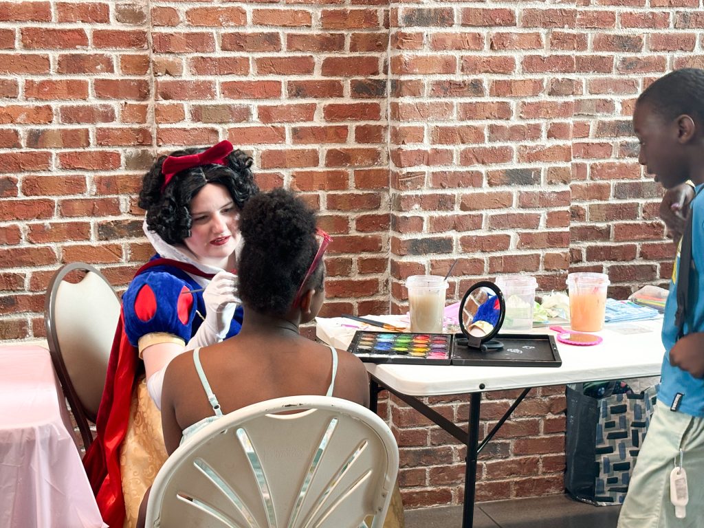 A woman dressed up as Disney's Snow White paints a girls face at a face painting station at a community event.