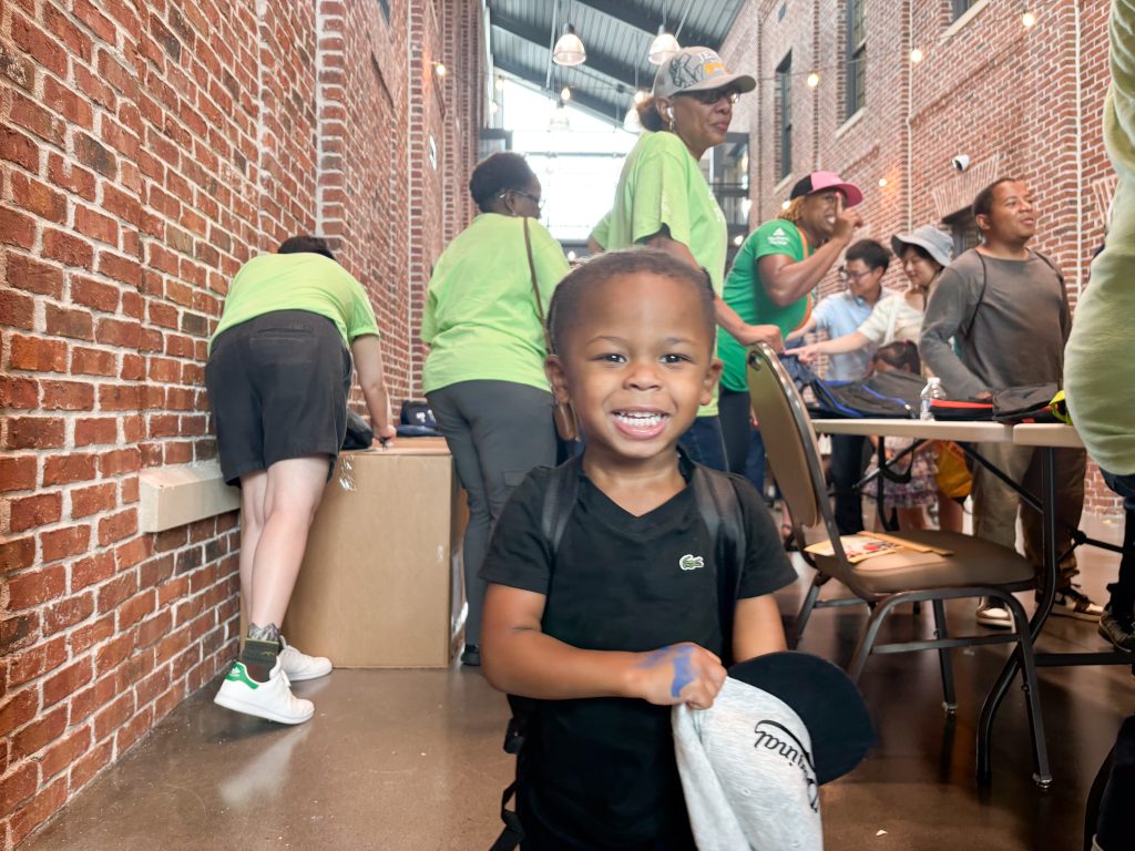 A young boy holds a ball cap and smiles while wearing a backpack filled with back-to-school items.