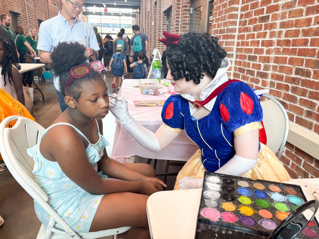 A woman dressed up as Disney's Snow White paints a girl's face at a face painting station at a community event.