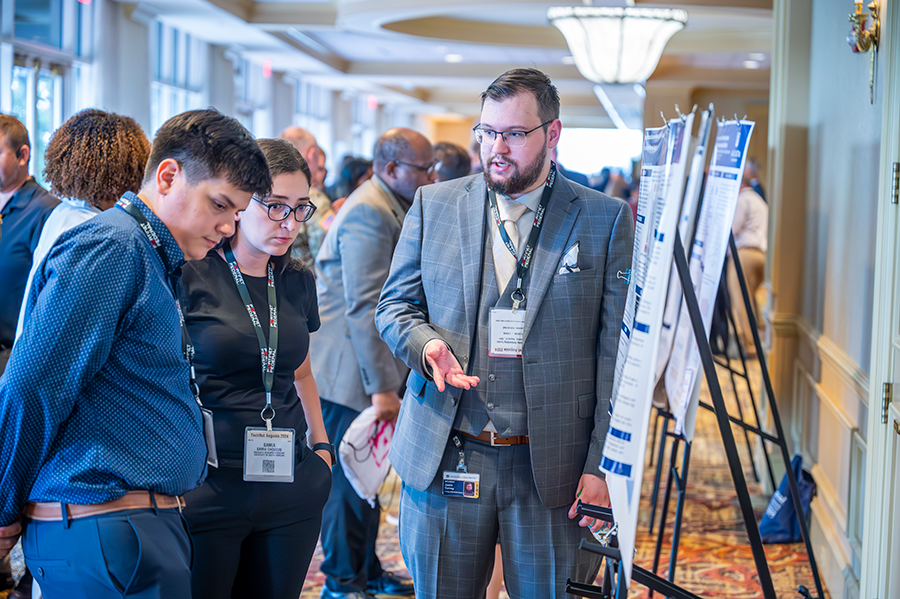 A college graduate student speaks with a man and a woman about information displayed on a large poster.