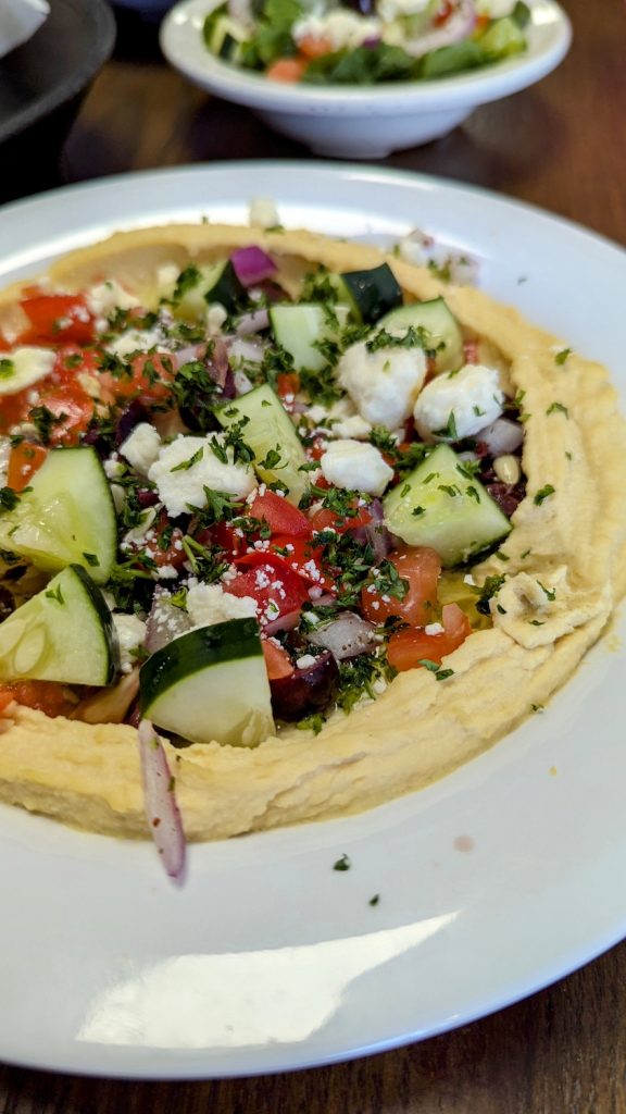 A dish of layered hummus with feta cheese, tomato, pico de gallo and other vegetables.