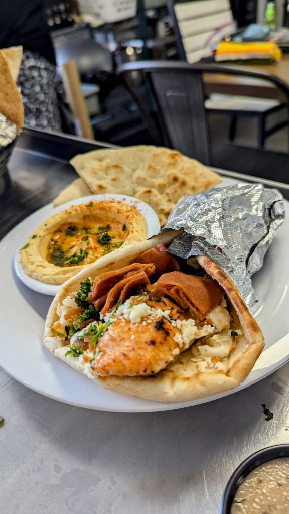 A small bowl of hummus sits next to a wrap containing grilled chicken, feta and yams.