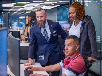 Three people are gathered around a computer screen with one man sitting at the keyboard. A man in a suit is leaning on the table the computer is sitting on while a woman looks on.