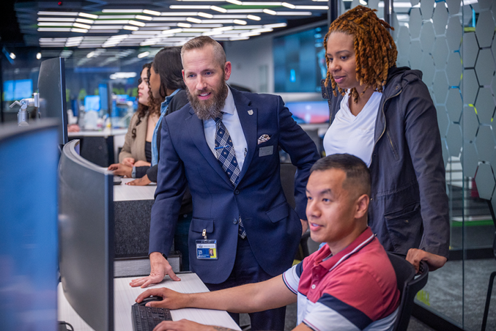 Three people are gathered around a computer screen with one man sitting at the keyboard. A man in a suit is leaning on the table the computer is sitting on while a woman looks on.