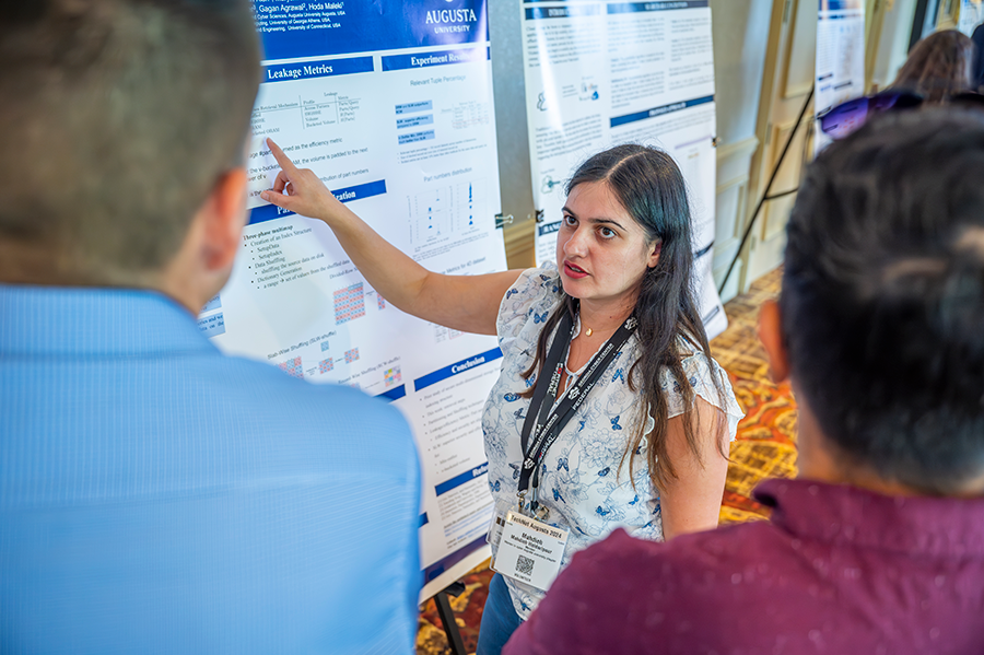 A woman points to information on a poster board that contains research information and talks to two people.