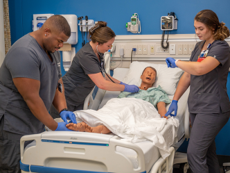 Three nursing students practice taking vital signs from a mannequin laying in a medical bed inside a lab set up to look like a hospital room.