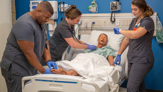 Three nursing students practice taking vital signs from a mannequin laying in a medical bed inside a lab set up to look like a hospital room.