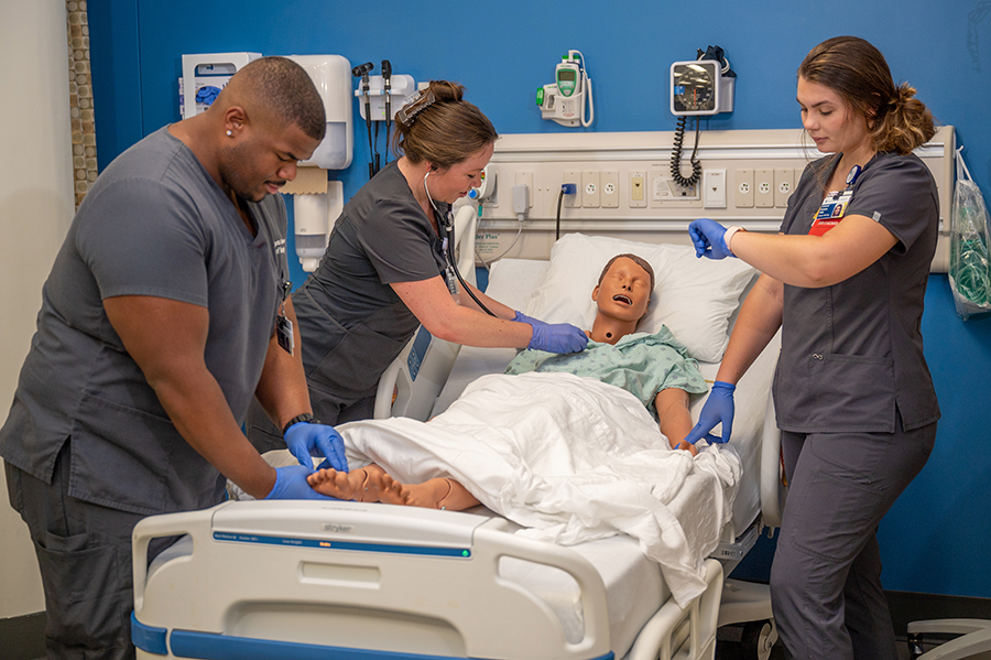 Three nursing students practice taking vital signs from a mannequin laying in a medical bed inside a lab set up to look like a hospital room.