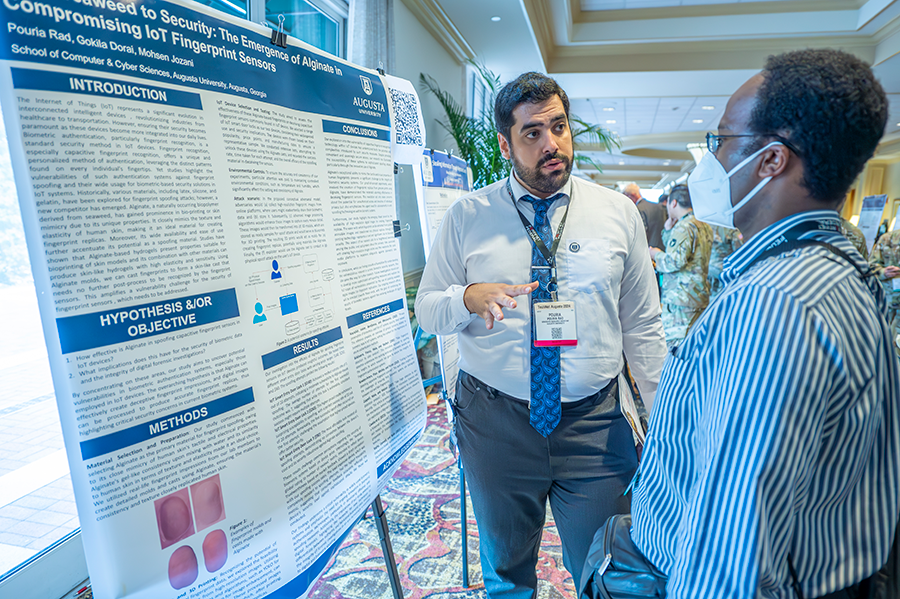 A man stands in front of a large poster with information about research and talks to another man.