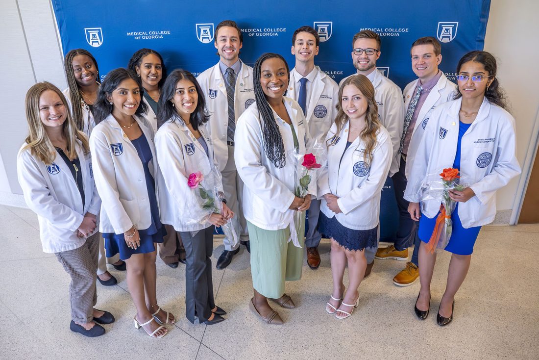 A group of 12 medical students in their medical-professional lab coats stand in front of a backdrop with logos for Augusta University and the Medical College of Georgia on it. The group includes eight women and four men. Three of the women are holding roses.