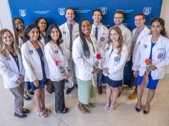 A group of 12 medical students in their medical-professional lab coats stand in front of a backdrop with logos for Augusta University and the Medical College of Georgia on it. The group includes eight women and four men. Three of the women are holding roses.