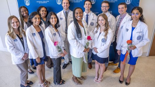 A group of 12 medical students in their medical-professional lab coats stand in front of a backdrop with logos for Augusta University and the Medical College of Georgia on it. The group includes eight women and four men. Three of the women are holding roses.