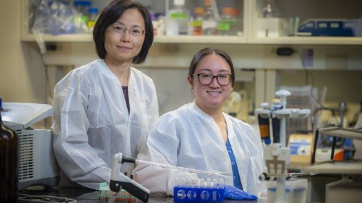 Two women in lab coats sit behind lab bench