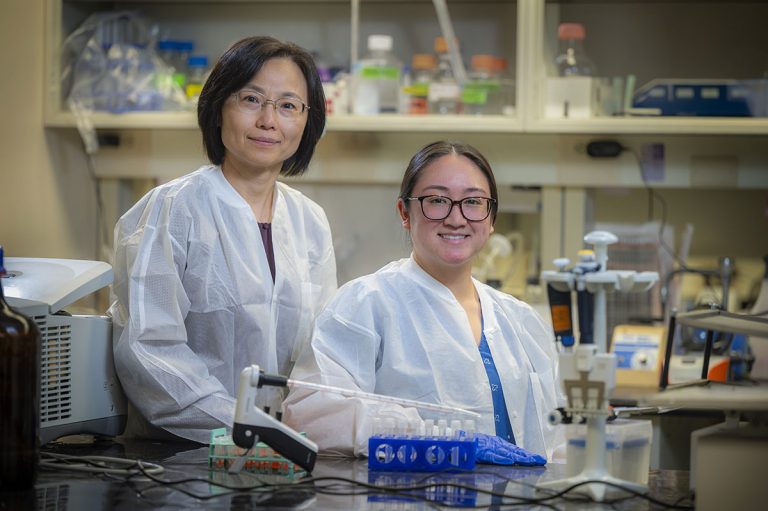 Two women in lab coats sit behind lab bench