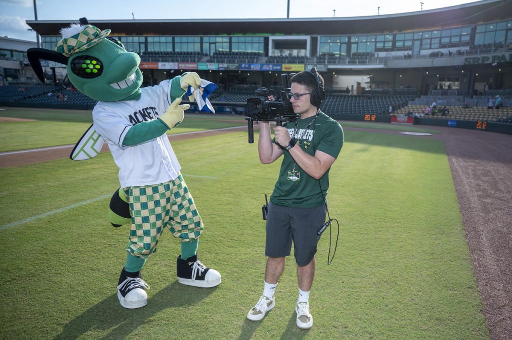 A student mans a camera as Auggie, the Augusta GreenJacket mascot, tries to distract him.