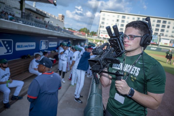 College student holds a camera outside a baseball dugout