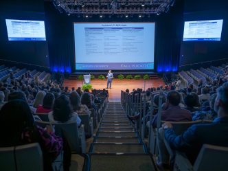 A man delivers a presentation on stage in front of an audience of hundreds.