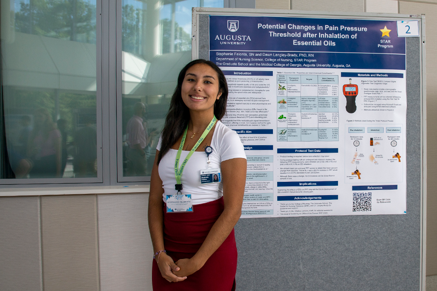 A college-aged woman stands in front of a presentation poster in a large atrium.