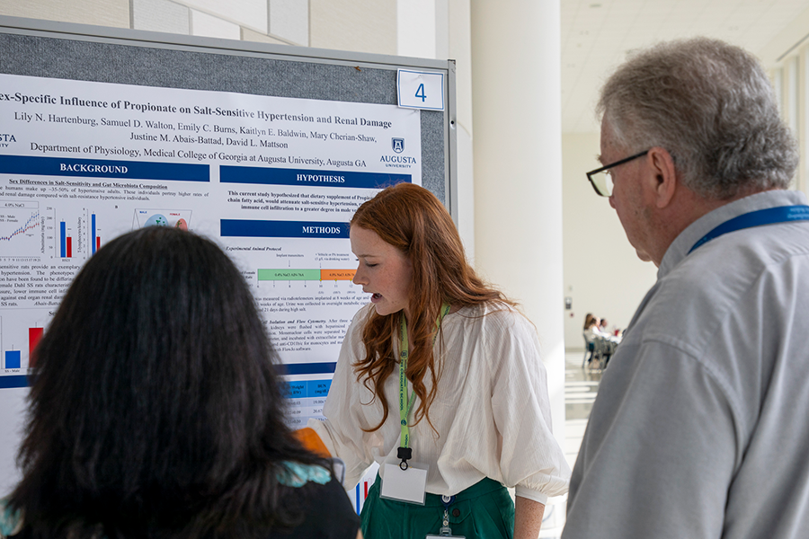 A group of people stand around a poster pinned to a moveable board. A college woman is talking to the group and pointing to pictures and graphs on the board.