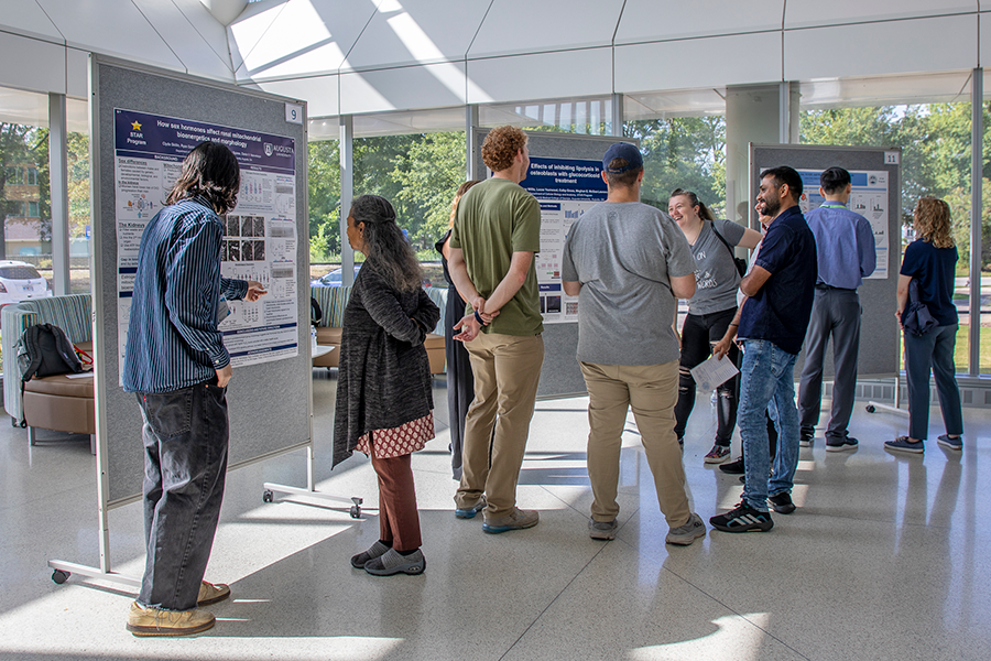 A large atrium is filled with people looking at rolling presentation boards. Each board has a poster on it detailing different types of research.