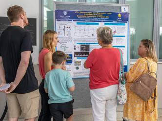 A group of people stand around a poster pinned to a moveable board. A college woman is talking to the group and pointing to pictures and graphs on the board.