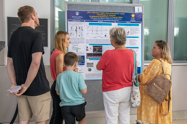 A group of people stand around a poster pinned to a moveable board. A college woman is talking to the group and pointing to pictures and graphs on the board.