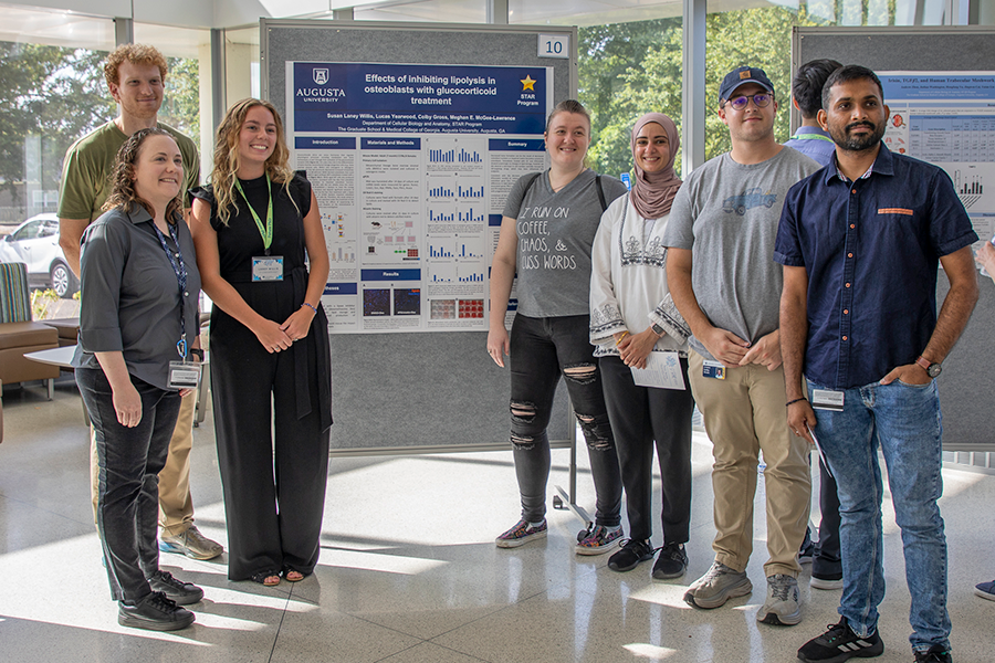 A group of college students and professors stand around a rolling board that has a poster on it. The poster features research, including graphs and photos.