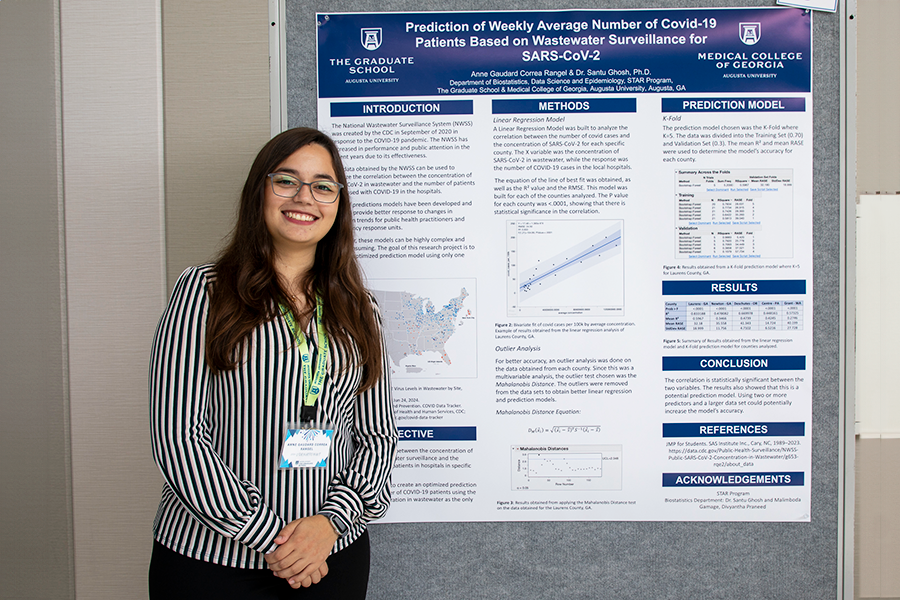 A college-aged woman stands in front of a presentation poster in a large atrium.