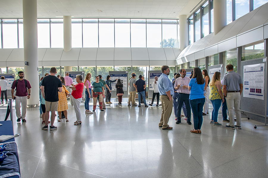 A large atrium is filled with people looking at rolling presentation boards. Each board has a poster on it detailing different types of research.