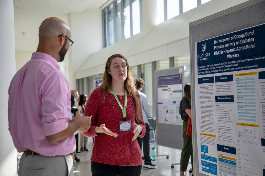 A college-aged woman stands in front of a presentation poster in a large atrium and speaks with a man about her research.