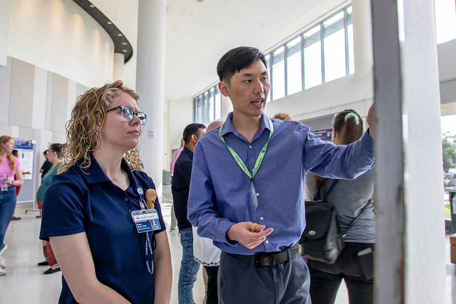 A male college student speaks with a female professor and points to a poster board while describing his research project.