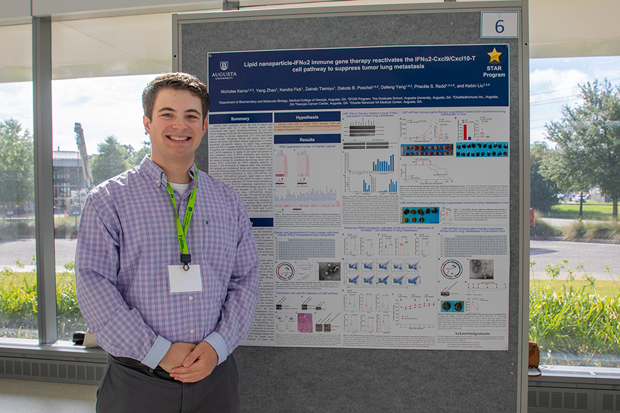 A college-aged man stands in front of a presentation poster in a large atrium.