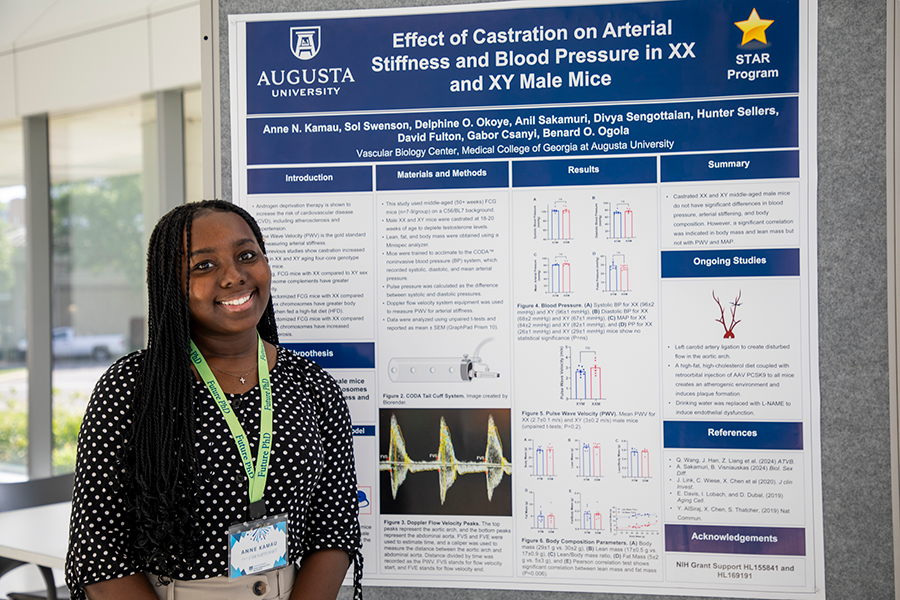 A college-aged woman stands in front of a presentation poster in a large atrium.