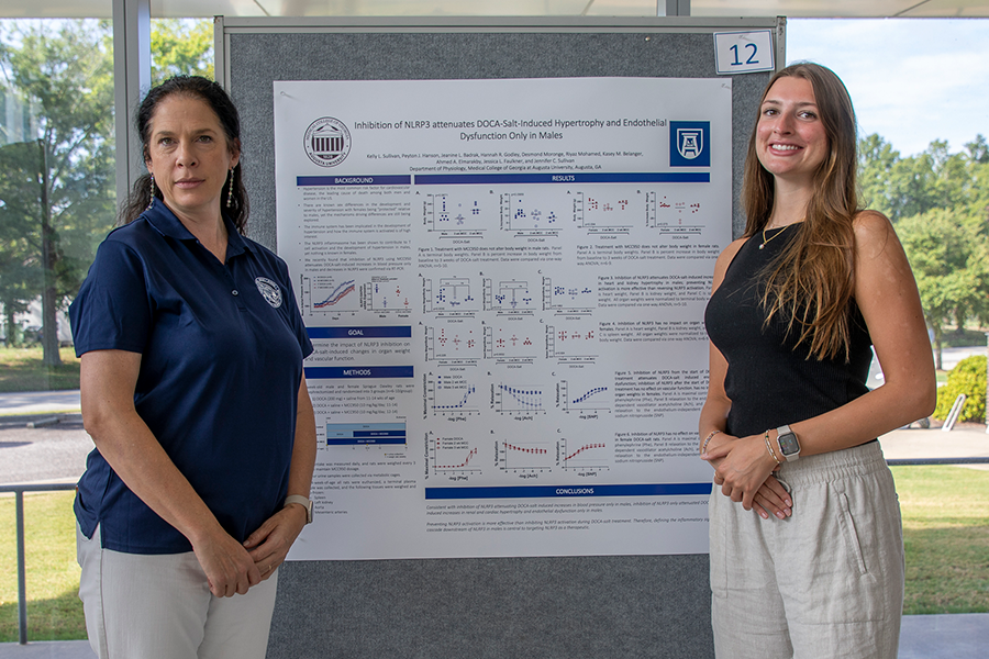 A college-aged woman and a college professor stand in front of a presentation poster in a large atrium.