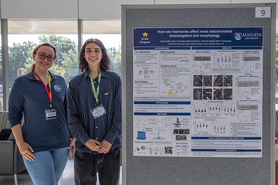 A college-aged man and a woman stand beside a presentation poster in a large atrium.