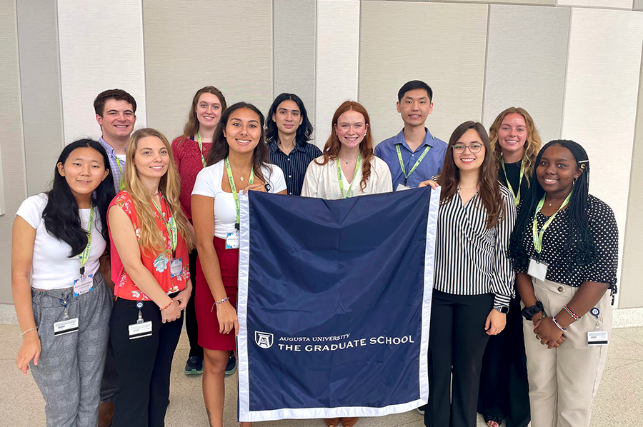 A group of 11 college-aged women and men stand around a flag that has "Augusta University The Graduate School" written on a solid background.