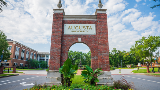 A brick archway with "Augusta University" carved into the front marks the entrance to a college campus.