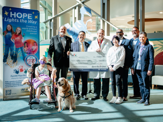 Six adults and two children, one in a wheelchair, stand in the lobby of a children's hospital. One adult is wearing a doctor's lab coat and is holding a large check.