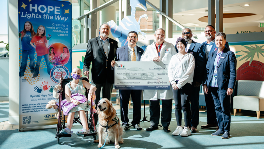 Six adults and two children, one in a wheelchair, stand in the lobby of a children's hospital. One adult is wearing a doctor's lab coat and is holding a large check.