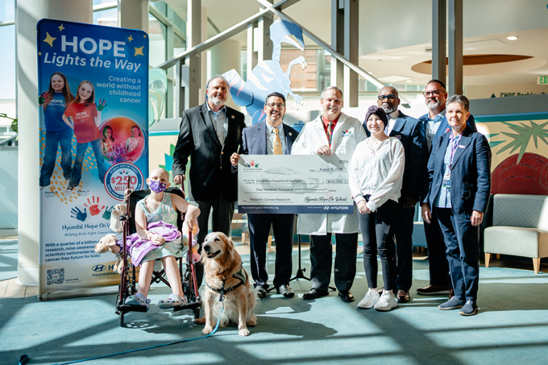 Six adults and two children, one in a wheelchair, stand in the lobby of a children's hospital. One adult is wearing a doctor's lab coat and is holding a large check.