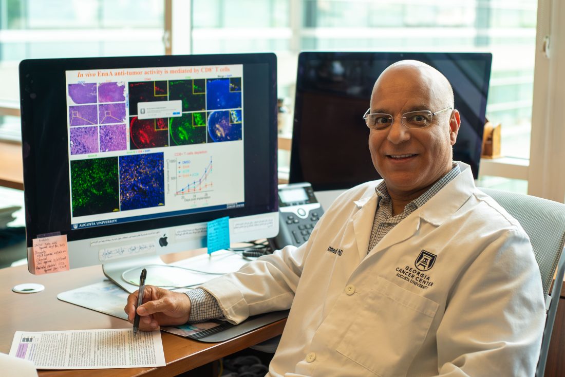 A man wearing a scientific lab coat sits at a computer station and writes notes on a pad of paper.