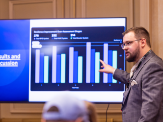 A man stands in front of a screen and presents to a large group about research depicted on the screen in the form of a bar graph.